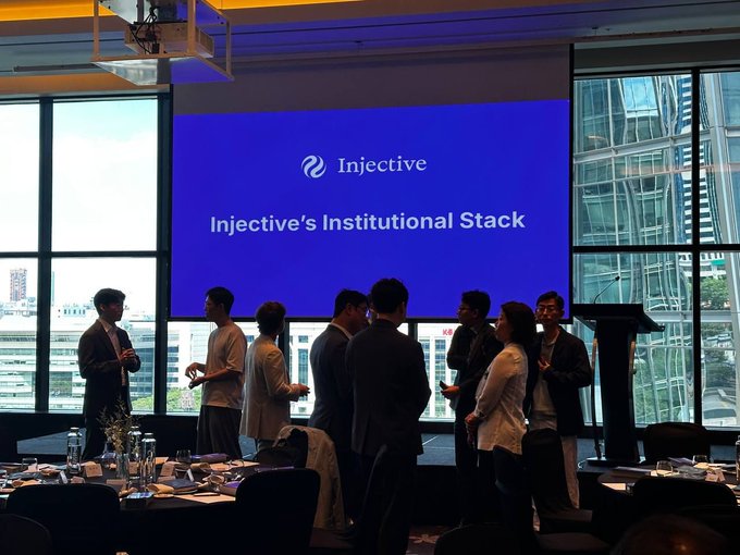 A group of people networking in a modern conference room with large windows, standing in front of a screen displaying "Injective" and "Injective's Institutional Stack".