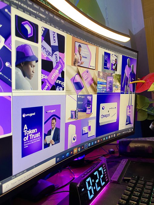 A curved monitor displaying a grid of 'unruggable' branded design mockups including 'A Token of Trust' marketing material, with a digital clock showing 19:22.