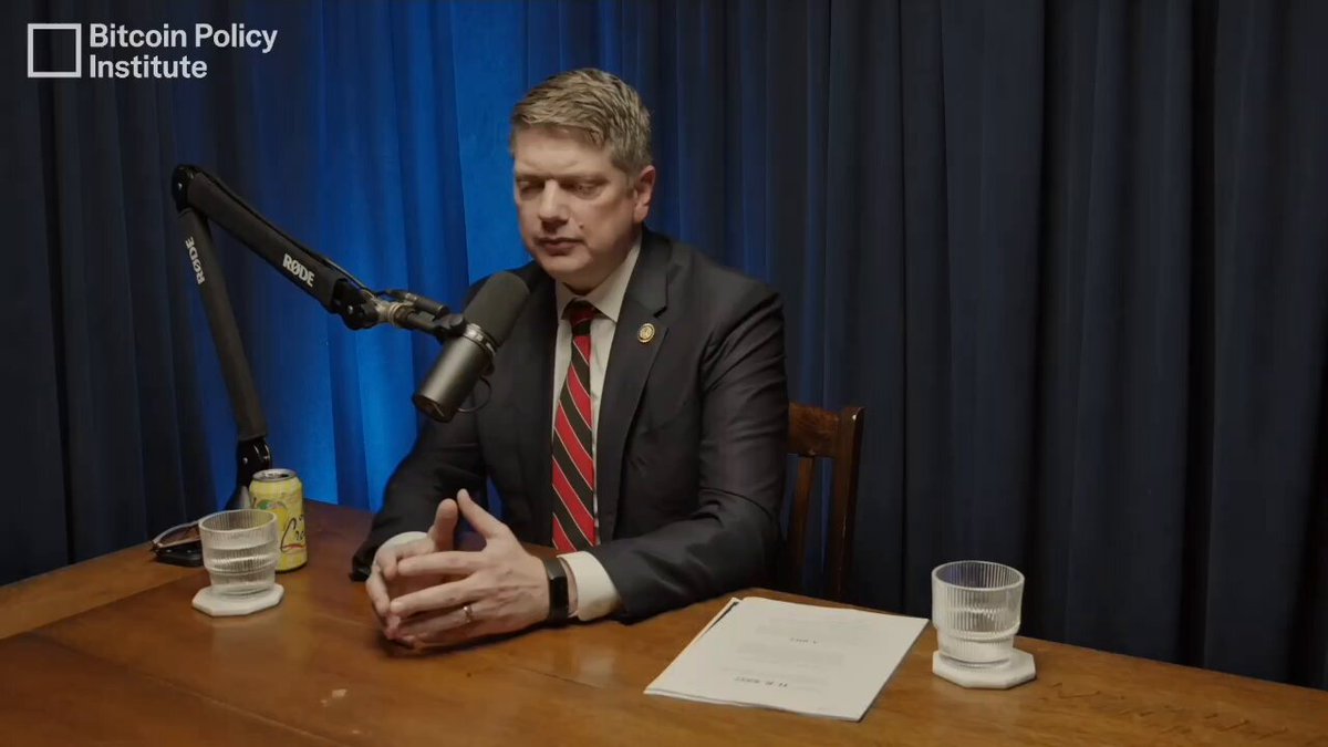 The image shows a man sitting at a desk wearing a suit and tie, holding a microphone with his hands crossed over them. The setting appears to be an office or studio environment, as there are two cups placed on the desk - one near the left side of the frame and another towards the right side. A TV screen is visible in the background, suggesting that this scene might be part of a live broadcast or interview. The image also contains text related to Bitcoin Policy Institute and XRP's path to U.S. government recognition for non-Bitcoin assets like XRP.