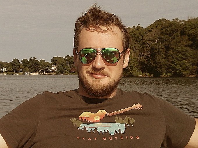 A bearded man wearing green-tinted aviator sunglasses and a brown 'PLAY OUTSIDE' graphic t-shirt takes a selfie by a lake surrounded by trees.