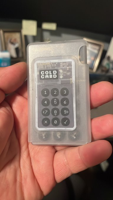 A hand holds a small "COLD CARD" hardware wallet featuring a numeric keypad (0-9, X, and checkmark), with "COLD CARD" displayed on its screen, representing the secure offline storage solution the poster wishes their friend would use instead of leaving Bitcoin on Coinbase. The image illustrates the tangible self-custody alternative that could prevent the heartbreak of losing access to one's crypto.