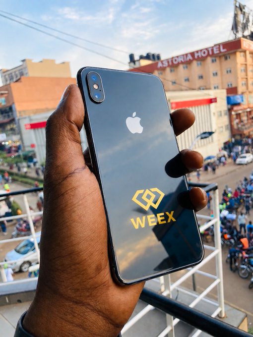 Photograph of a hand holding an iPhone with a 'WEEX' sticker on the back, with the 'ASTORIA HOTEL' visible in the urban background.