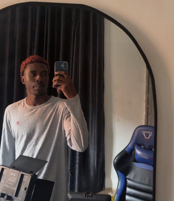 A mirror selfie of a young man with reddish-orange hair wearing a white long-sleeve shirt, standing in a room with dark curtains and a blue gaming chair.