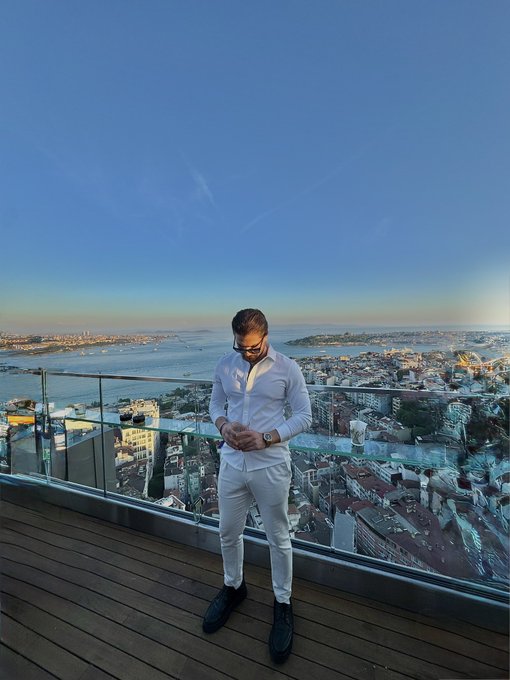 A man in an all-white outfit standing on a rooftop balcony overlooking a coastal city at sunset.