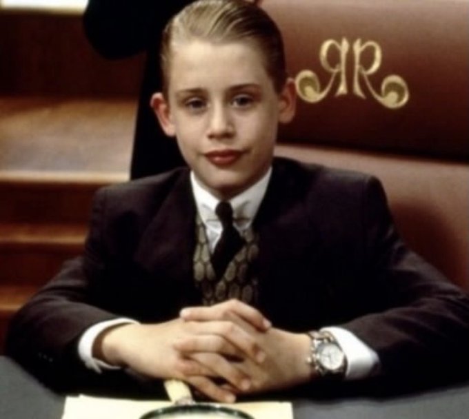 A young boy with slicked-back hair in a suit and tie, sitting at a table with his hands folded and wearing a wristwatch, with the gold initials 'AR' visible on the booth behind him.