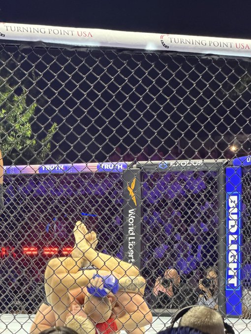 The image shows a man wrestling behind a chain-link fence, with another wrestler holding him down. The scene takes place at an indoor arena where there are people watching the match. A crowd of spectators can be seen around the ring, observing the action between the two wrestlers. In addition to the main event, there is also a beer advertisement visible in the background.