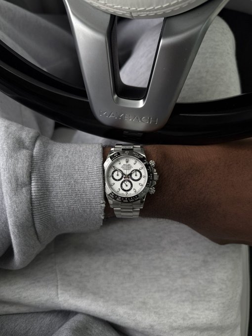 Wrist shot of a Rolex Cosmograph Daytona on a steel Oyster bracelet, resting on a grey sweatshirt beside a Maybach steering wheel. "GOOD MORNING MONDAY" — pairing luxury timekeeping with the start of a focused weekend.