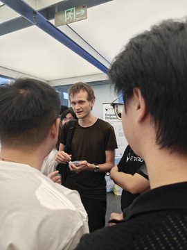 Several men gather in a crowded indoor area, including a central man in a dark brown T-shirt holding a phone.