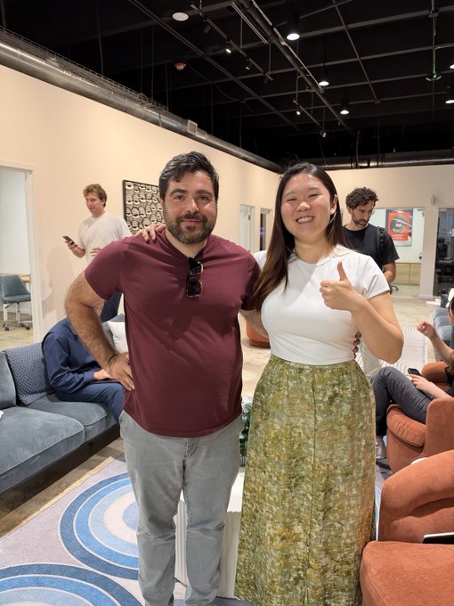 Two people posing together at an indoor gathering; a man in a maroon shirt stands beside a woman in a white top and patterned green skirt who is giving a thumbs up.