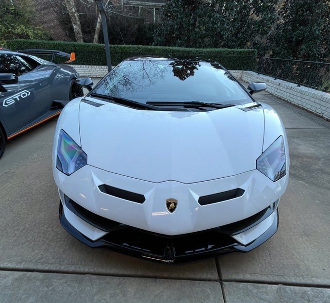 A white Lamborghini Aventador is parked in a driveway next to a gray sports car with "GTO" branding visible on its side. The image relates to the caption's joke about choosing between multiple "fake" Lamborghinis, showcasing an impressive exotic car collection with the ticker $TBB mentioned.