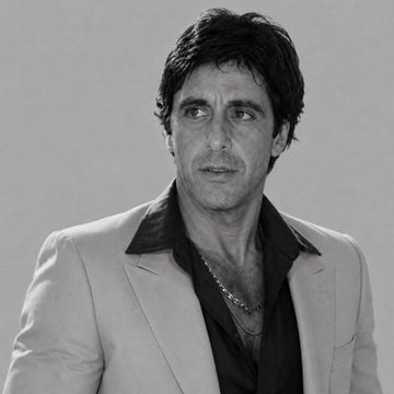 A black-and-white studio portrait shows a dark-haired man in a light-colored suit jacket over a dark open-collar shirt, looking slightly off-camera.