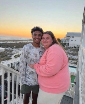 A couple smiling at the camera on a balcony, with the ocean and sunset visible behind them.
