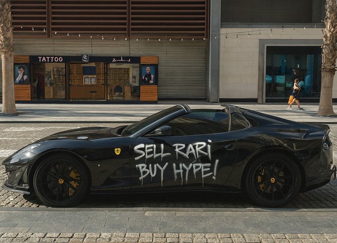 A black Ferrari sports car parked on a street with 'SELL RARI BUY HYPE!' written in white graffiti on the door, with a 'TATTOO' shop in the background.
