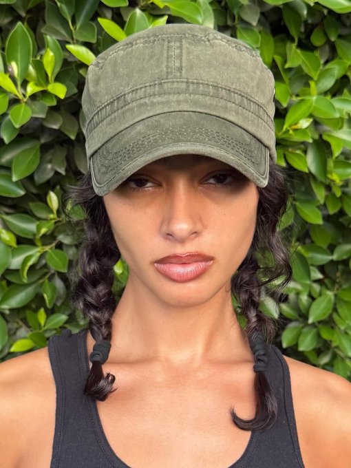 A woman with dark braided hair wearing a faded green military-style cap and black tank top stands in front of a leafy green hedge.