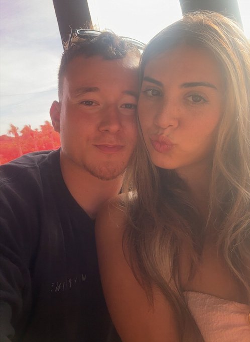 A close-up selfie of a young couple; a man in a dark shirt on the left and a woman with long brown hair making a kissy face on the right, taken outdoors.