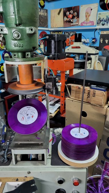 Purple vinyl records being pressed by a vintage green industrial machine, with labels reading "ANGLO" and "SIDE A."

The image shows a record pressing facility in action, illustrating the caption's narrative of the song "Juro que..." finally being pressed onto vinyl after its creation at the Songcamp organized by @sweetman_eth.