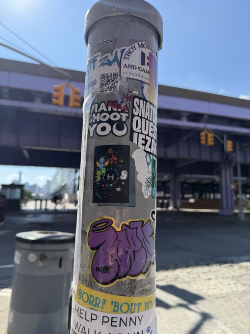 A sticker-covered metal pole on a city street featuring various decals including one that reads 'SNATCH MY QUEST!' and another saying 'HELP PENNY'.