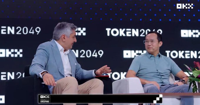Two men seated on stage at the 'TOKEN 2049' conference, one in a light blue blazer gesturing while speaking to the other in a teal polo shirt.