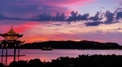 A vibrant sunset scene over a calm body of water featuring a silhouetted traditional pavilion on the left and a small boat in the distance, with pink and purple clouds.