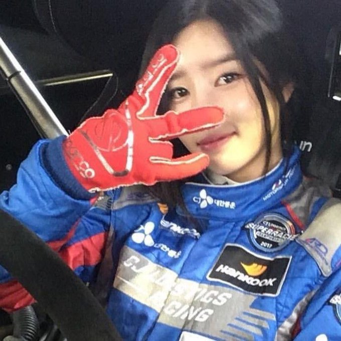 A smiling woman in a blue racing suit and red Sparco gloves poses inside a vehicle, with 'CJ Logistics Racing' and 'Hankook' branding visible.