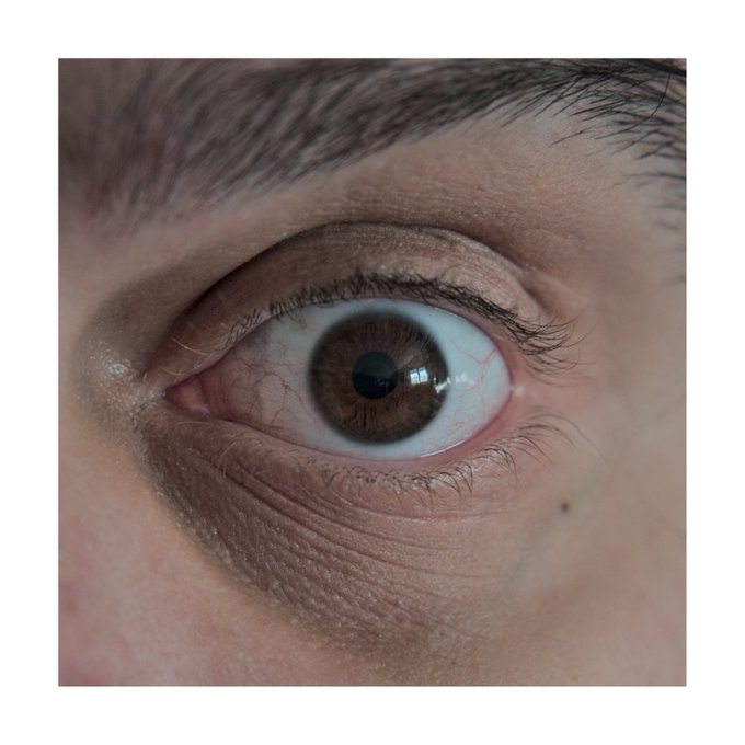 A close-up photograph of a hazel eye with visible blood vessels, framed by a dark eyebrow and surrounding skin, serving as the artwork for an NFT listing on objkt.com. The image's intimate, vulnerable gaze pairs with the author's tongue-in-cheek "poem" about the non-fungible joy value of their digital art.