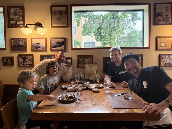 A group of men and children enjoy a meal at Destefano's Steakhouse, with the window displaying "DeStefanos" in reverse lettering and the wall covered in framed photos and memorabilia. The lively gathering captures the casual, fun atmosphere referenced in the post about hanging out with Web3 friends and family at the DeStefanos clubhouse.