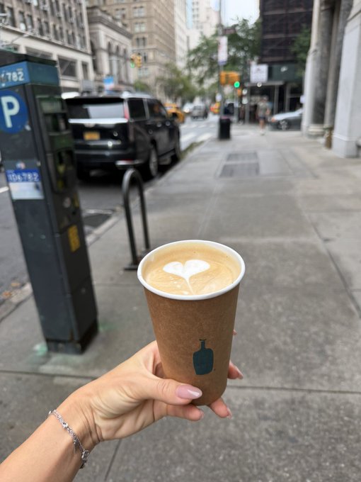 A hand with pink manicured nails holds a brown paper coffee cup with a blue bottle logo containing a latte with heart-shaped foam art, set against an urban street scene with a parking meter labeled 'P 10679-2' and city traffic.