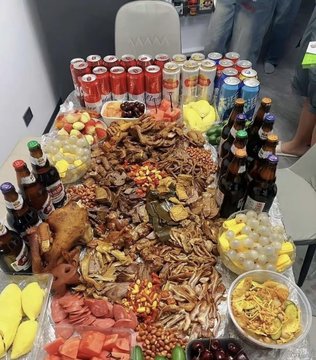 A table laden with a feast of assorted nuts, dried fruits, cold cuts, fruits, beer bottles, and soda cans.