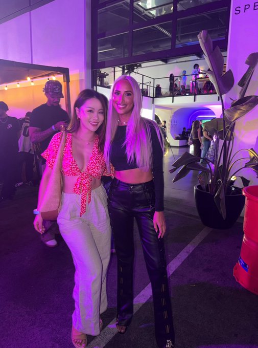 Two women pose together at an outdoor evening event with purple lighting; one wears a red patterned top with light pants, while the other wears a black crop top and leather pants.
