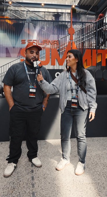 Two people being interviewed at a Solana Summit event, with the man in a Solana cap and lanyard holding a microphone, and the woman in a striped blazer, standing in front of a 'SUMMIT' banner.