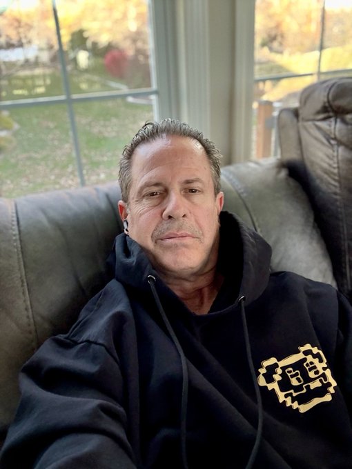 A middle-aged man with short, graying hair sits on a gray leather couch, wearing a black hoodie featuring a yellow pixelated graphic.