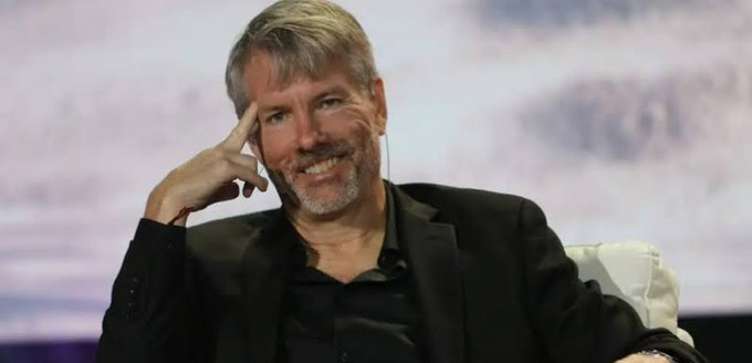 A smiling man with gray hair and a beard, wearing a black blazer over a black shirt, sits on stage with his hand resting against his temple. The image likely depicts Michael Saylor, founder of MicroStrategy (now branded as Strategy), who is known for championing the company's Bitcoin-focused treasury strategy referenced in the accompanying post about raising $467M through stock sales to boost its USD Reserve.