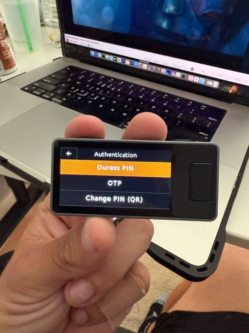 A hand holds a hardware wallet displaying an authentication menu with options for "Duress PIN," "OTP," and "Change PIN (QR)," illustrating the security measures crypto holders rely on amid rising physical threats. The "Duress PIN" feature appears highlighted, a safeguard that allows users to create a decoy wallet under coercion—a response to the surge in kidnappings and attacks on Bitcoin holders referenced in the post.