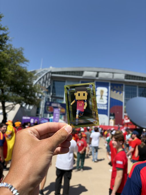 A hand holding up a small trading card featuring a blocky character with the text 'LUKAKU 9', displayed at a stadium with crowds of fans in red jerseys, likely at a World Cup event.
