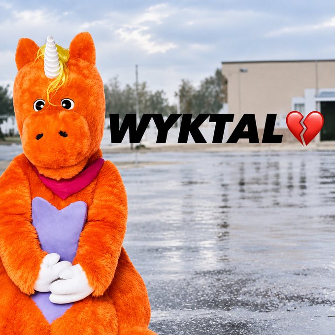 An orange unicorn mascot character sitting with hands clasped, holding a purple heart, set against a parking lot background, with the text 'WYKTAL' and a broken heart emoji.