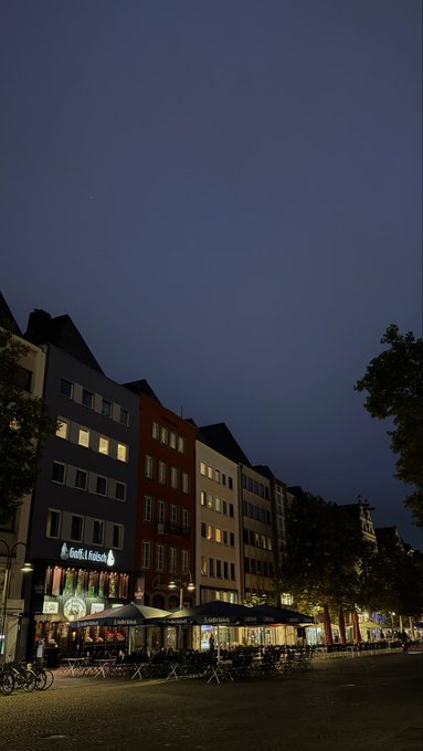 A twilight street scene shows illuminated storefronts and outdoor café seating, with a prominent "Grill & Fritte..." restaurant sign visible among the row of buildings under a dark blue sky. The peaceful, quiet city atmosphere complements the post's "goodnight" sentiment, suggesting a calm evening before tomorrow's web3 events.