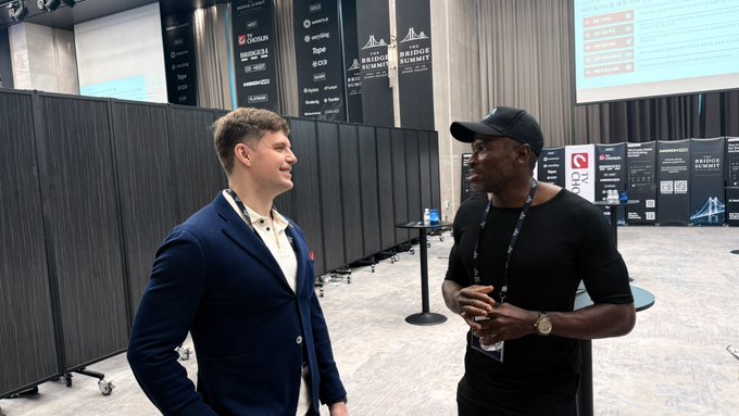 Two men having a conversation at an indoor event; one wears a blue blazer and the other a black t-shirt and cap, with "National Alumi" banners visible in the background.