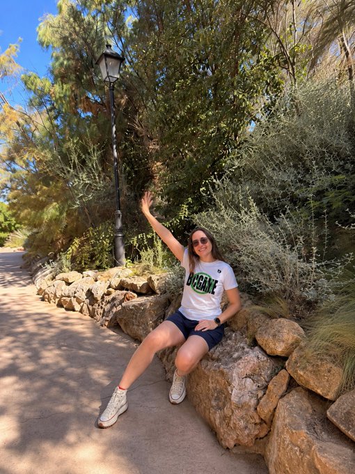 A woman wearing sunglasses and a 'BOGAN' t-shirt sits on a rocky ledge along a sunlit garden path, waving at the camera.