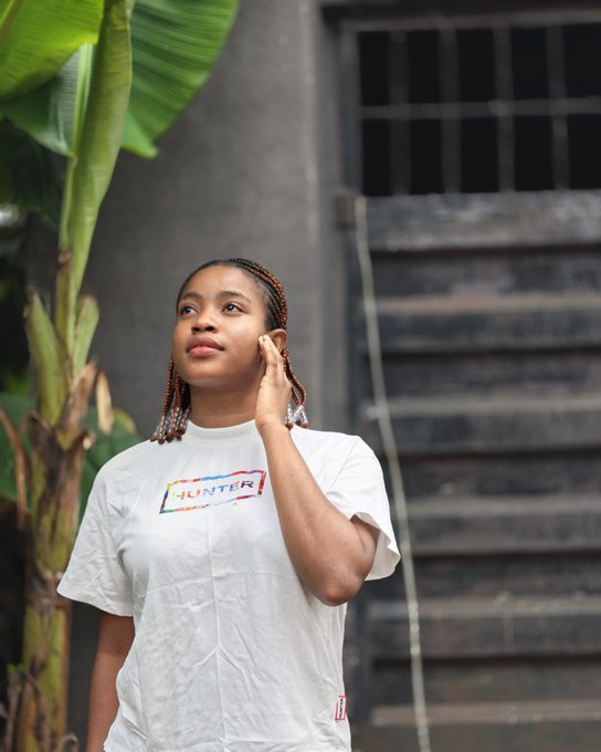 A portrait of a young woman with braided hair wearing a white 'HUNTER' branded t-shirt, posing in front of a building with stairs and tropical green foliage in the background.