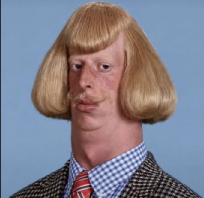A portrait of a man with a distinctive blonde bowl haircut, mustache, checkered shirt, striped tie, and tweed jacket against a blue background.