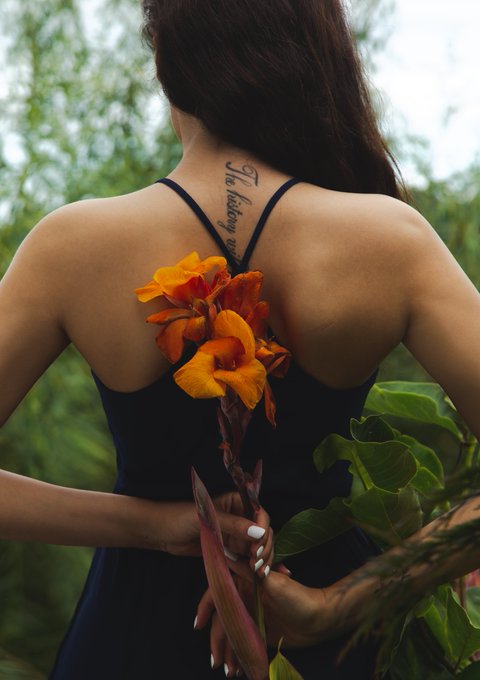 A photograph of a woman's back showing a tattoo reading 'She believed she could' between her shoulder blades, as she holds orange flowers (canna lilies) behind her back while wearing a dark dress.