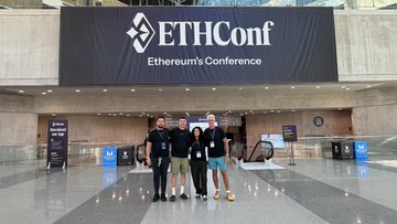 The image shows a group of four people standing together in front of a large banner that says "ETHCONF" written in white letters. The group appears to be posing for a photo at an event, possibly an ethercon's conference or meeting. They are all dressed similarly and seem to be enjoying the occasion.