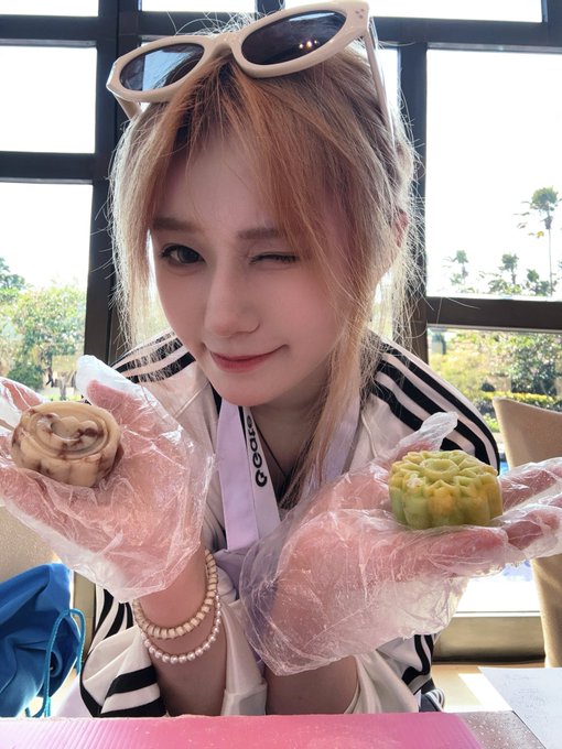 A young woman winks while holding two pastries wearing plastic gloves, with sunglasses perched on her head.