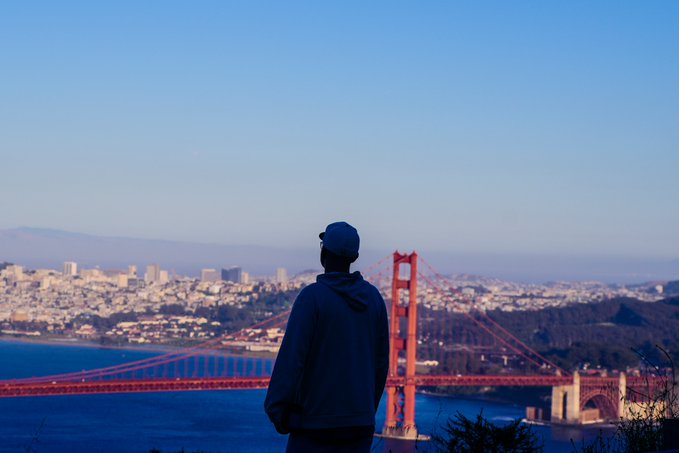A person wearing a blue hoodie and cap stands looking out at the Golden Gate Bridge with the San Francisco skyline in the background under a clear sky.