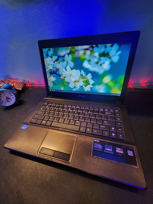 An ASUS laptop open on a desk displaying a cherry blossom wallpaper, with an analog alarm clock to the left and colorful LED string lights in the background.