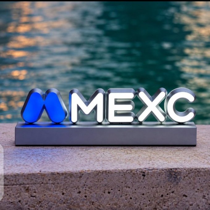 An illuminated MEXC cryptocurrency exchange logo sign rests on a stone ledge with rippling blue water in the background.