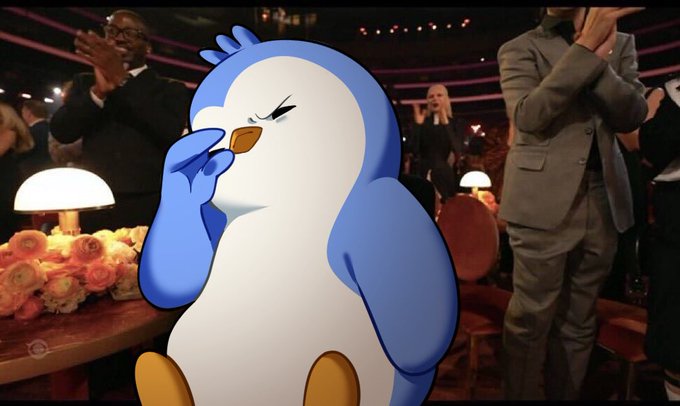 An animated blue penguin character with closed eyes and a hand to its face in a dismissive gesture, set against a background of people clapping at what appears to be an awards show or event.