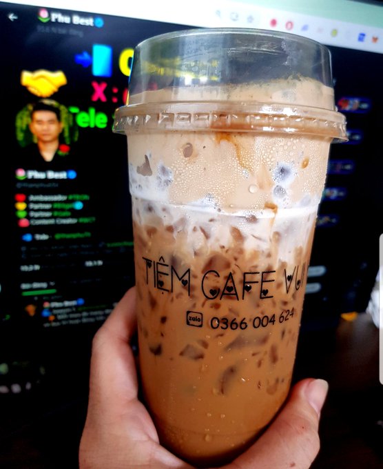 A hand holding a plastic cup of iced coffee with the text 'TIỆM CAFE' and the phone number '0366 004 624' printed on it, with a computer screen displaying a social media or chat page visible in the background.