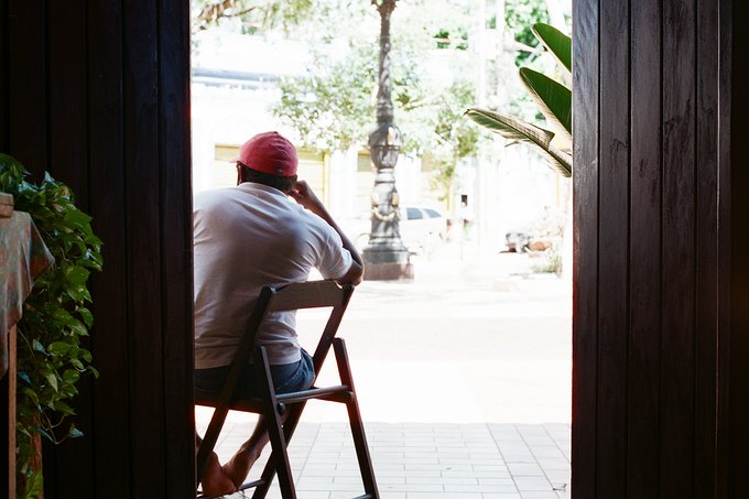 A man in a white shirt and red cap sits on a folding chair in a doorway, gazing out onto a sunlit street with a vintage lamppost and parked cars, embodying the contemplative stillness of "lost in memories during the fading days of spring."