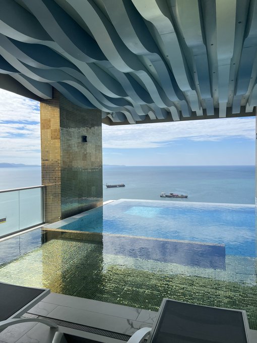 A luxurious rooftop infinity pool overlooks the ocean with cargo ships on the horizon, framed by a striking wavy blue-and-white architectural ceiling and a gold-tiled wall.